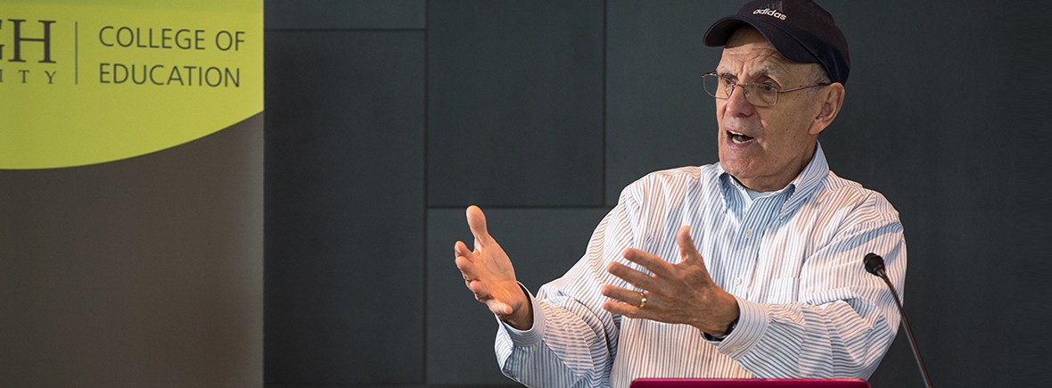 A photo of Zirkel wearing a navy baseball cap giving a lecture from a lectern with a microphone.